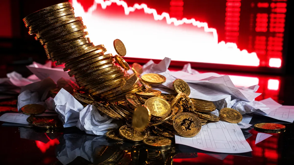 赤く下落する株価チャートを背景に、崩れかけたビットコイン硬貨の山と、クシャクシャに丸められた大量の書類